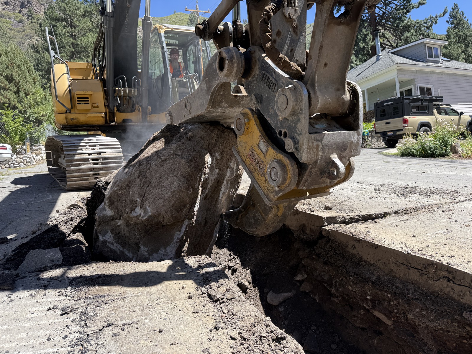 Excavator pulling a boulder out of a trench cut through pavement, operator in hard hat and hi-vis vest in the cab