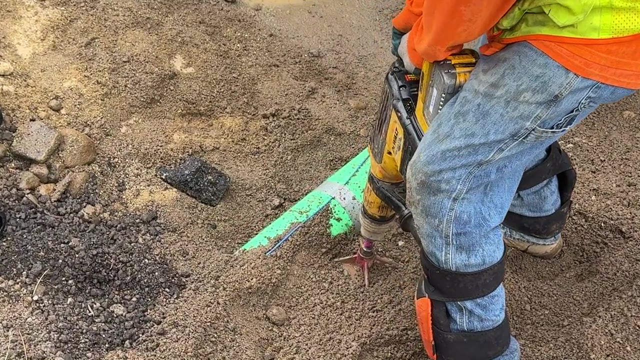 Worker in hi-vis running a DeWalt SDS max rotary hammer with a spade bit into gravelly ground beside green utility-marking paint
