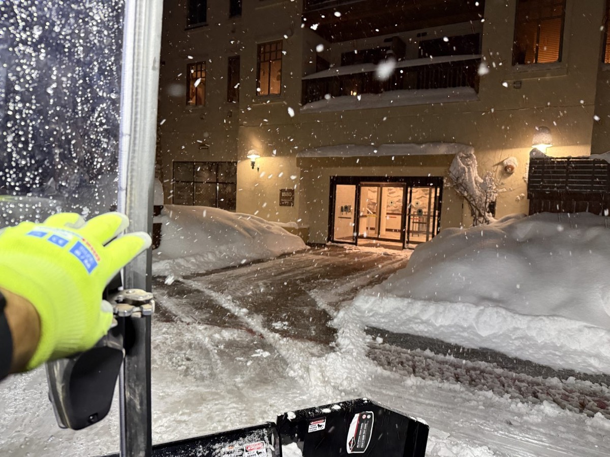 Operator's view from the cab at night — heavy snowfall burying a lit village entrance, hi-vis glove on the controls