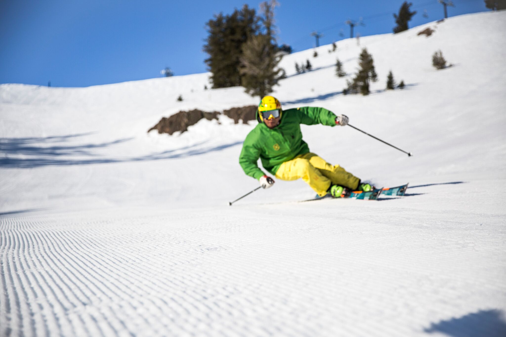 Errol carving a groomed run on a bluebird day in the green and yellow Jamaica kit, chairlift and rock outcrop behind
