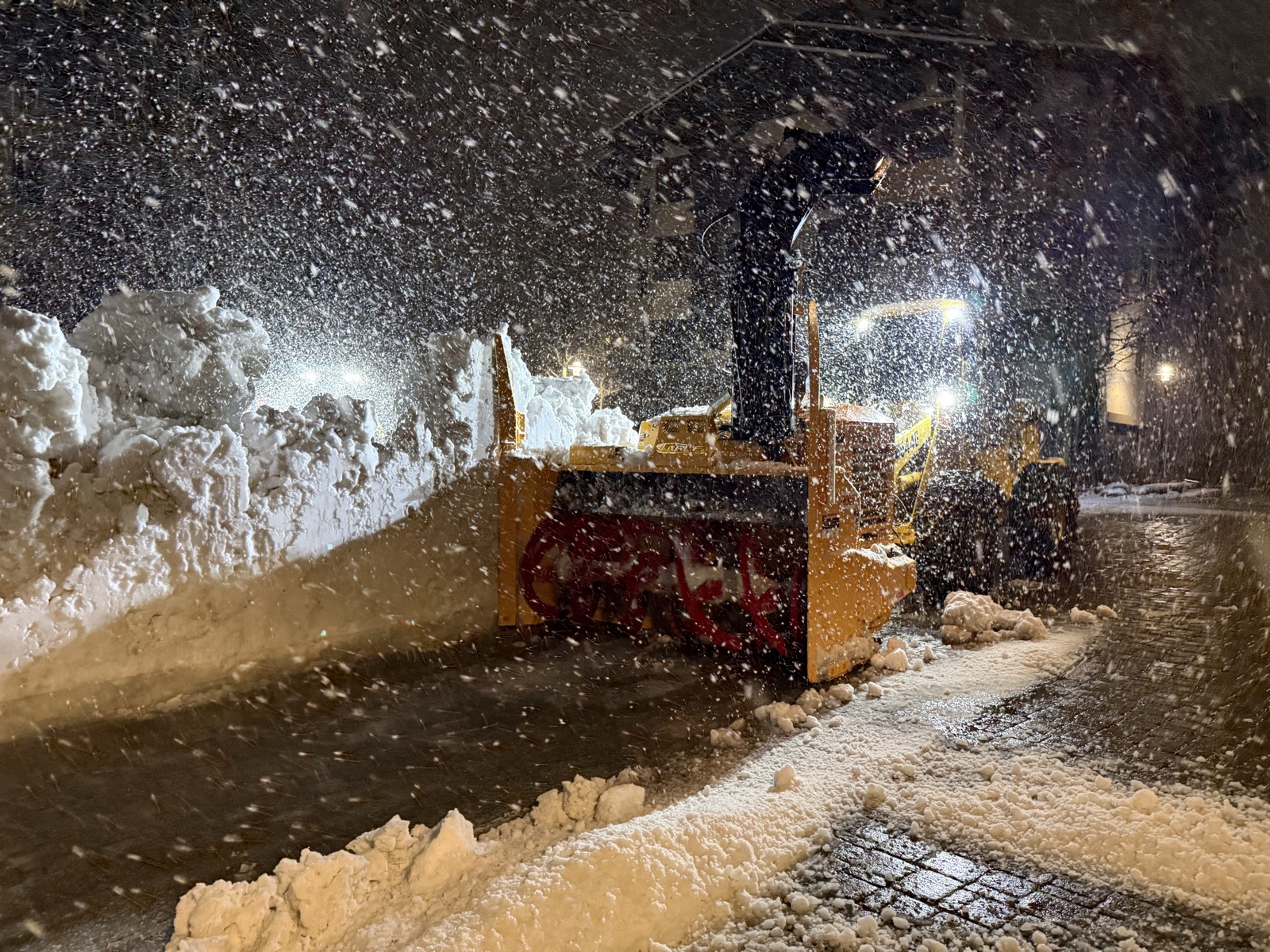 Loader-mounted snowblower running at night in heavy snowfall beside a tall snowbank