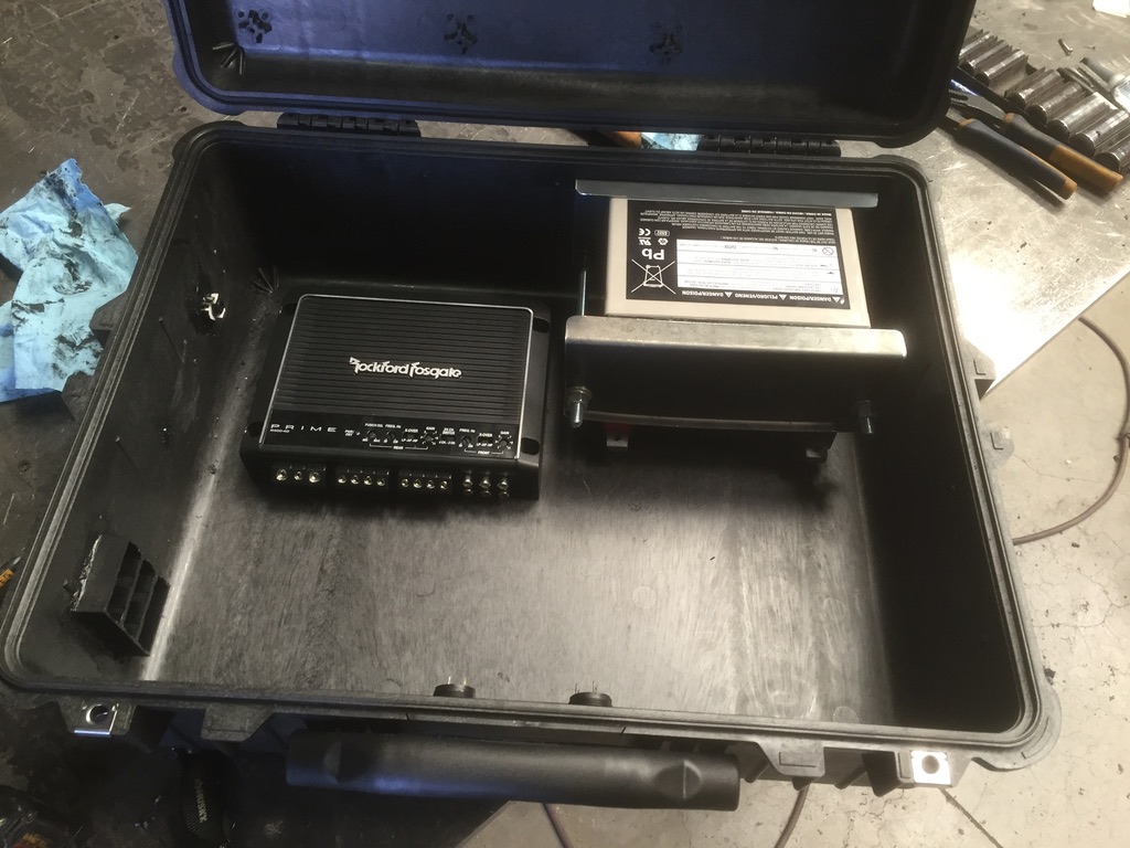 Open Pelican 1600 during the build: Rockford Fosgate Prime amplifier sitting on the left of the case floor, battery on the right held down by a welded steel cradle, with one of the louvered vents visible on the interior wall