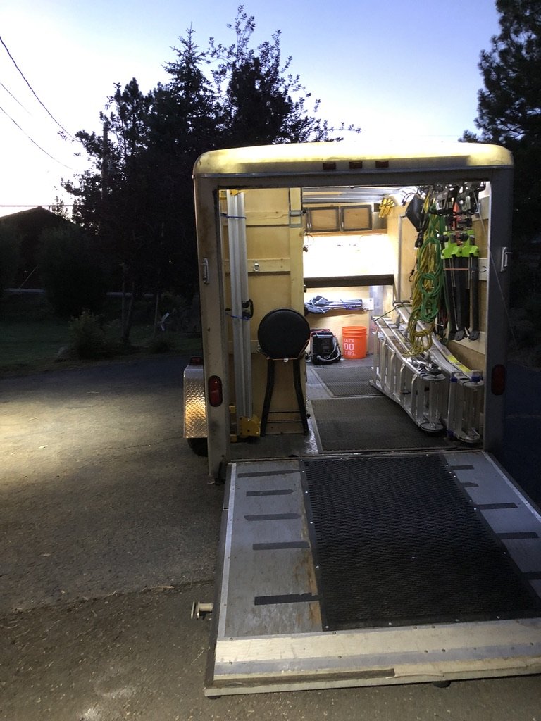 DIY mobile carpentry trailer at dusk, ramp open and interior lit — extension cords and ladders stowed on the wall