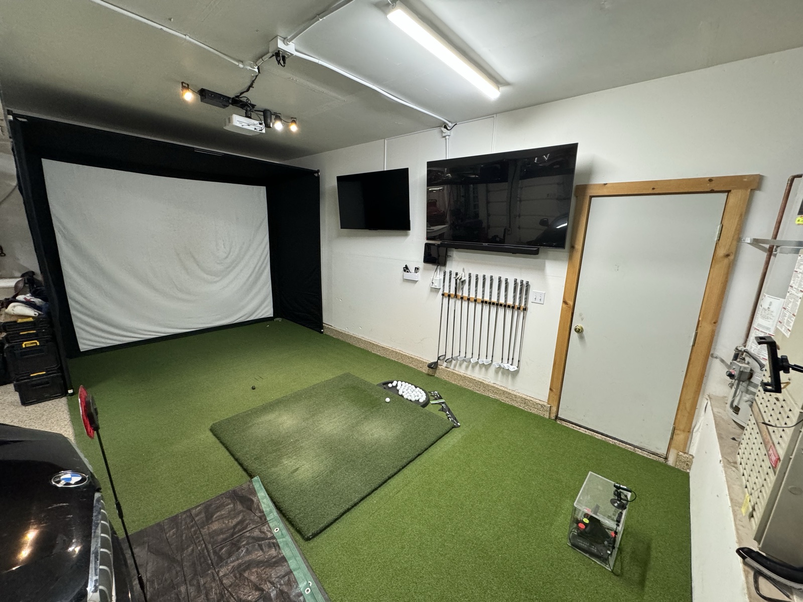 Golf simulator with retractable impact screen rolled down, putting green turf, wall-mounted TV, 14 clubs on wall rack, projector ceiling-mounted