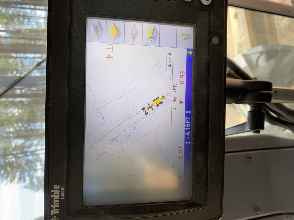 Trimble CB460 grade control display in an equipment cab showing cut and fill numbers against a digital design surface, pine trees visible through the windshield