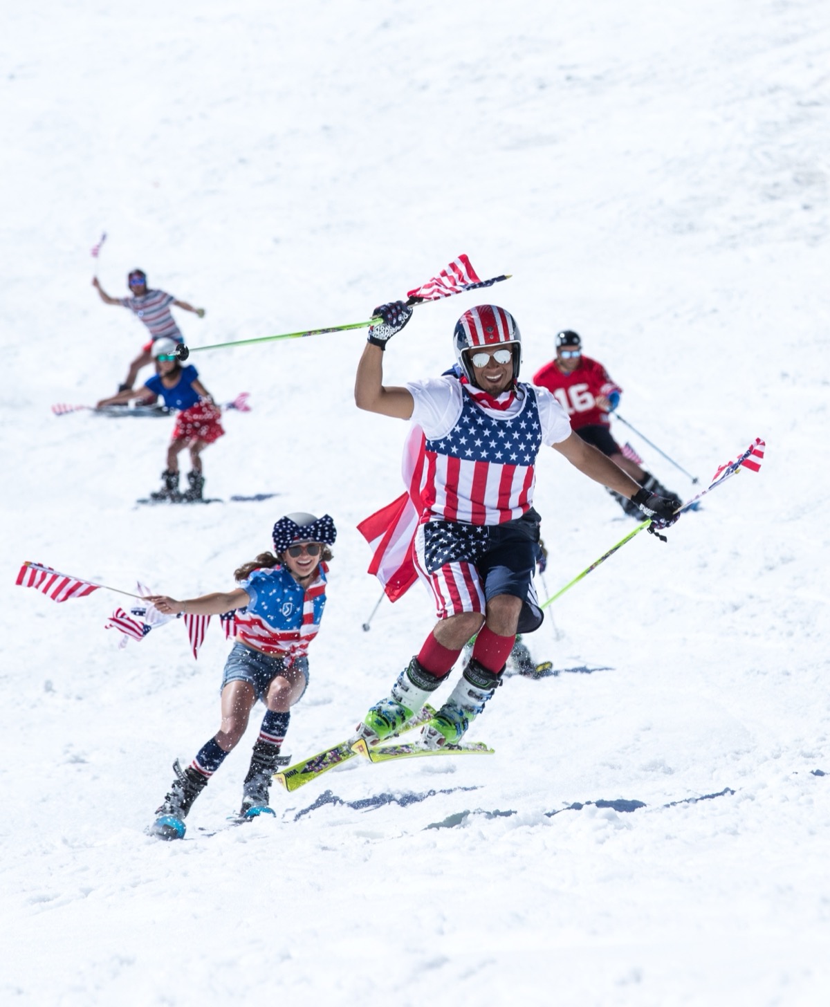 Skiers and snowboarders in American flag gear, flags in hand, charging down the hill at a Fourth of July ski event